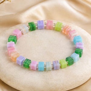 Può includere: Un braccialetto composto da piccole perline cubiche multicolori nelle tonalità del verde, rosa, blu, viola e arancione, separate da minuscole perline distanziatrici sferiche trasparenti, disposte in un motivo circolare su una superficie liscia e chiara.