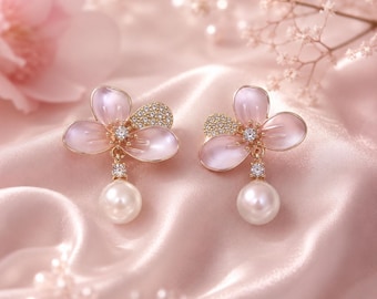 May include: A pair of gold-tone earrings featuring three light pink, petal-shaped elements arranged in a flower design. One petal is encrusted with small clear stones. A single clear stone connects the flower to a dangling white pearl drop.