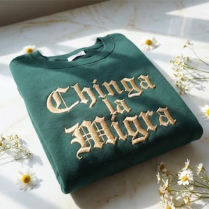 Könnte beinhalten: Ein gefaltetes, dunkelgrünes Sweatshirt mit dem gestickten Text "Chinga la Migra" in goldener, gotischer Schrift. Das Sweatshirt ist von kleinen weißen und gelben Gänseblümchen umgeben.