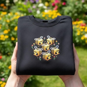 以下が含まれることがあります： 小さな花に囲まれた、黄色と黒の6匹の蜂のピクセル化されたデザインが特徴の黒いスウェットシャツ。 蜂は円形に配置されています。 スウェットシャツは折りたたまれ、ぼやけた花の背景の前に保持されています。