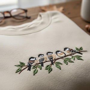 May include: A cream-coloured crew neck sweatshirt featuring an embroidered design of five small birds on a branch with green leaves. The birds have black, white, and brown markings. The sweatshirt is made of a soft, textured fabric.