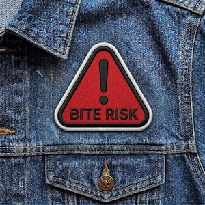 May include: A red triangular patch with a black exclamation point and the words "BITE RISK" in black lettering. The patch has a white border and is sewn onto a blue denim jacket.
