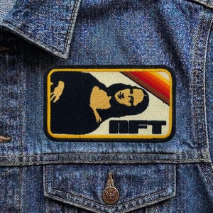May include: A rectangular patch with a retro design featuring a woman's silhouette and the word "NFT" in bold letters. The patch has a black border and is set against a denim jacket.