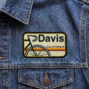 May include: A rectangular patch with the word "Davis" in black letters and a bicycle graphic. The patch has a cream-colored background with a bicycle wheel design in orange and yellow. The word "CALIFORNIA" is below the bicycle wheel.