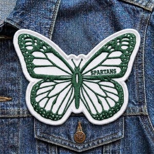 May include: A white and green butterfly patch with the word "SPARTANS" embroidered on the wing. The patch is sewn onto a denim jacket. The butterfly design features intricate details and a white border.