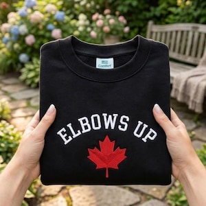 May include: Black sweatshirt with the text "ELBOWS UP" in white, arched above a red maple leaf. The sweatshirt has a crew neck and is held by hands. The tag reads "Comfort".