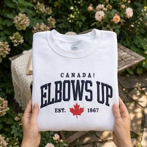 May include: A white sweatshirt with the text "CANADA! ELBOWS UP EST. 1867" in navy blue lettering and a red maple leaf. The sweatshirt is held up by a person's hands, with a blurred background of greenery and flowers.