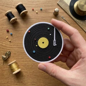 May include: A black embroidered patch in the shape of a vinyl record with a yellow center and white trim. The patch has a white needle and colorful bead accents. Other sewing notions, including spools of thread, are in the background.
