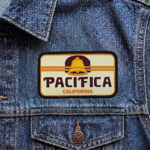 May include: A rectangular patch with the word "PACIFICA" in burgundy letters, and "CALIFORNIA" in smaller gold letters. The patch features a gold bell design and is set against a cream background with gold borders. The patch is sewn onto a denim jacket.