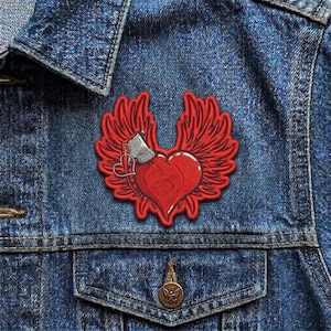 May include: A red heart-shaped patch with red wings and a small silver-colored axe. The patch is on a blue denim jacket. The heart has a small heart-shaped charm attached to it.