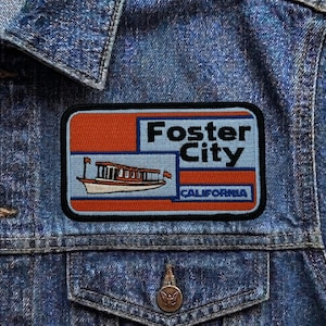 May include: A rectangular patch with the words "Foster City California" and a boat illustration. The patch has a blue background with orange and white accents, and is sewn onto a denim jacket.