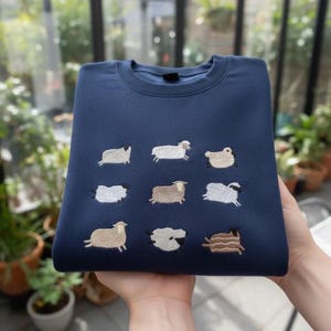 Puede incluir: Una sudadera azul marino con un diseño bordado de nueve ovejas de varios colores. Las ovejas están representadas en una postura de carrera o pastoreo. La sudadera está doblada y sostenida en las manos.