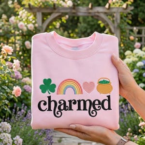 Könnte beinhalten: Hellrosa Sweatshirt mit dem Wort "charmed" in schwarzer Schrift. Das Design umfasst ein grünes Kleeblatt, einen Regenbogen, ein rosa Herz und einen Goldtopf. Das Sweatshirt ist gefaltet und wird gehalten.