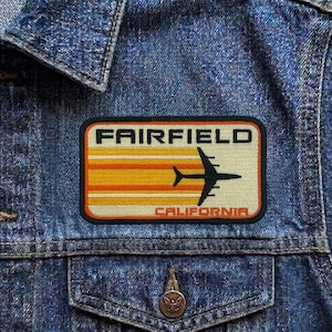 May include: A rectangular patch with the text "FAIRFIELD CALIFORNIA" and a graphic of a black airplane. The patch has a cream background with orange and yellow horizontal stripes. The patch is sewn onto a denim jacket.