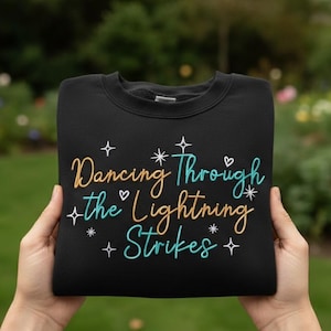 Könnte beinhalten: Schwarzes Sweatshirt mit Rundhalsausschnitt und dem Text "Dancing Through the Lightning Strikes" in goldener, türkisfarbener und weißer Schrift. Das Design umfasst Sterne und Herz-Akzente. Das Sweatshirt wird von Händen gehalten.