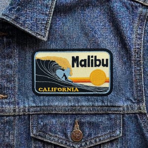 May include: A rectangular patch with the word "Malibu" and "California" on a denim jacket. The patch features a wave with a surfer, a setting sun, and a cream-colored background.