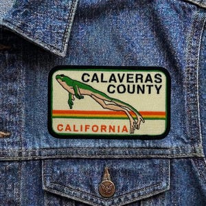 May include: A rectangular patch featuring a green frog with the text "CALAVERAS COUNTY CALIFORNIA". The patch has a cream background with green, orange, and yellow stripes. The patch is sewn onto a blue denim jacket.