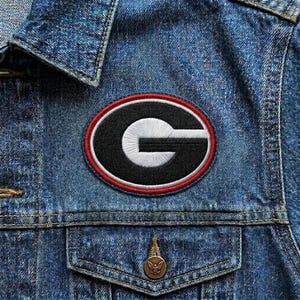 May include: Embroidered patch featuring the University of Georgia Bulldogs logo, a white "G" on a black oval with a red and white border. The patch is sewn onto a blue denim jacket.