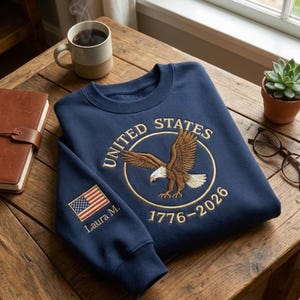 May include: Navy blue sweatshirt with an embroidered eagle design inside a circle that reads "UNITED STATES 1776-2026." The sleeve features an American flag and the name "Laura M." A coffee mug, notebook, and plant are also visible.