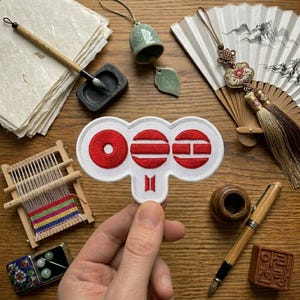May include: A white patch with three red circles and a red horizontal bar design, with the BTS logo below. The patch is held by a hand, surrounded by a wooden table, a fan, a bell, a brush, and other Asian-inspired items.