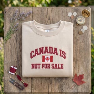 May include: Beige sweatshirt with the text "CANADA IS NOT FOR SALE" in burgundy lettering and a Canadian flag graphic. The sweatshirt is folded on a wooden surface, with a sprig of lavender, coins, and a compass nearby.