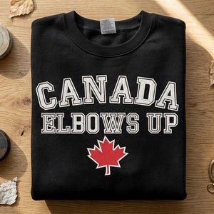 May include: Black sweatshirt with the words "CANADA ELBOWS UP" in white block letters and a red maple leaf graphic. The sweatshirt is folded on a wooden surface, with hockey pucks and a compass nearby.