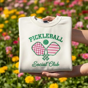 May include: Cream-colored sweatshirt with green embroidered text and graphics. The text reads "PICKLEBALL Social Club" above a pickleball and two paddles. One paddle has a pink and white checkered design, the other has pink and white stripes.