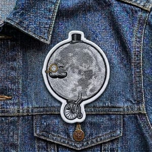 May include: A circular patch featuring a detailed illustration of the moon with a monocle, mustache, and top hat, riding a penny-farthing bicycle. The patch is embroidered with gray and white thread and has a white border. The patch is affixed to a denim jacket.