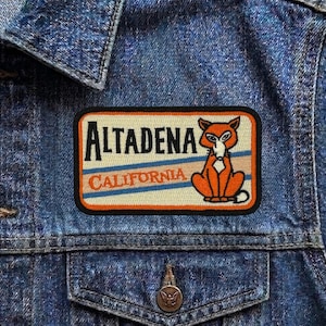 May include: A rectangular patch with the text "ALTADENA CALIFORNIA" and a cartoon fox illustration. The patch has a cream background with orange and blue stripes, and a black border. The patch is sewn onto a denim jacket.