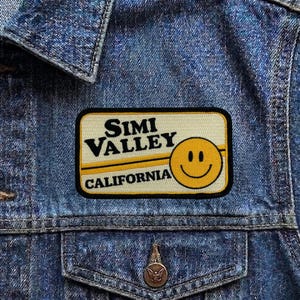May include: A rectangular patch with the words "Simi Valley California" and a yellow smiley face. The patch is on a blue denim jacket. The text is in a black font with a yellow and black border.