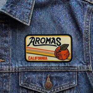 May include: A rectangular patch with the word "AROMAS" in a script font, and "CALIFORNIA" in a sans-serif font. The patch features an orange with a green leaf and horizontal stripes in shades of orange, brown, and yellow.
