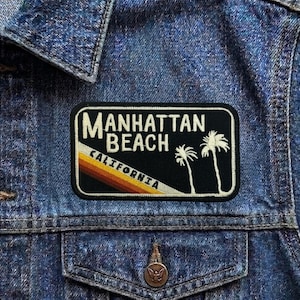 May include: A rectangular patch with the words "Manhattan Beach California" in white lettering. The patch features a black background with two white palm trees and a diagonal stripe of orange, yellow, and brown.