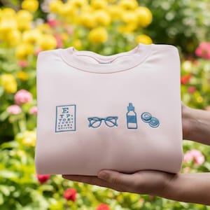 May include: A light pink sweatshirt with embroidered blue eye care symbols. The design includes an eye chart, eyeglasses, a bottle of eye drops, and contact lenses. The sweatshirt is held in front of a colorful floral background.