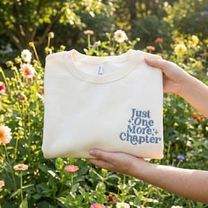 May include: Cream-colored sweatshirt with the text "Just One More Chapter" in blue. The crew neck sweatshirt is folded and held in front of a floral background.