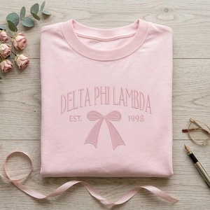 Könnte beinhalten: Hellrosa Sweatshirt mit gesticktem Text "DELTA PHI LAMBDA EST. 1998" und einem Schleifendesign. Das Sweatshirt ist ordentlich auf einer Holzoberfläche gefaltet, mit einem Band, Blumen, Brille und Stift.