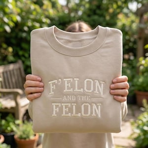 May include: A beige sweatshirt with the text "F'ELON AND THE FELON" in a bold, off-white font. The sweatshirt is held up in front of a person, with a blurred background of greenery and a wooden bench.