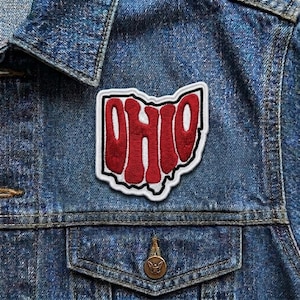 May include: Embroidered patch in the shape of Ohio with the word "OHIO" in red, outlined in white and black. The patch is affixed to a blue denim jacket, with a pocket and a button visible.