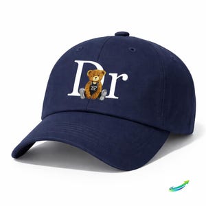 May include: Navy blue baseball cap with the letters "Dr" in white. A brown teddy bear wearing a black jersey with the number "02" is centered between the letters. The cap has a curved brim and a button on top.