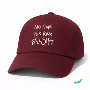 Pode incluir: Boné de beisebol bordô com a frase "NO TIME FOR YOUR BULLSHIT" em branco. O boné tem uma aba curva e um botão na parte superior. O chapéu é de cor sólida e tem um estilo casual.