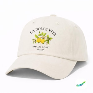 Op de afbeelding: Crèmekleurige baseballpet met de tekst "LA DOLCE VITA" boven een citroentakillustratie. Daaronder staat de tekst "AMALFI COAST ITALIA". De pet heeft een gebogen klep en een knoop aan de bovenkant.