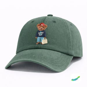 May include: A green baseball cap with a curved brim. The cap features an embroidered design of a teddy bear wearing a red cap, a navy blue sweater with the text "LUCKY GUY", and blue jeans. The bear is holding a white bag.
