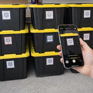 May include: Black storage bins with yellow lids are stacked in a garage. Each bin has a white label with a QR code. A smartphone is held up to scan a QR code on one of the bins.