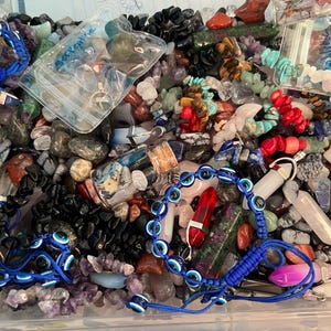 May include: A collection of various gemstones and jewelry, including bracelets with blue evil eye beads, crystals, and loose stones in shades of blue, red, black, and purple. Some items are in clear plastic bags.
