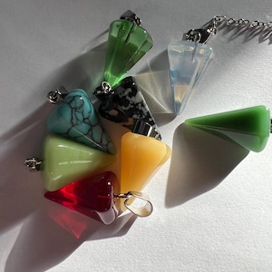 May include: A collection of colorful crystal pendulums in various shapes and colors, including green, red, yellow, and turquoise. Each pendulum has a silver-colored metal loop for hanging. The pendulums are arranged on a white surface.