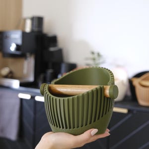 Könnte beinhalten: Olivgrüne Kaffeesatz-Abschlagbox mit Holzstange. Die Abschlagbox hat ein geripptes Design und wird in einer Hand gehalten. Im Hintergrund ist eine Kaffeemaschine zu sehen.