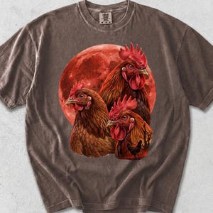 May include: A dark brown short-sleeve t-shirt featuring a graphic print of three chickens positioned in front of a large, textured red moon. The design includes detailed feathers and red combs on the birds, set against the circular lunar background.