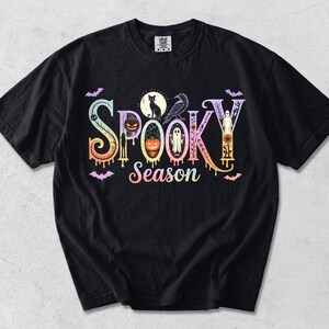 以下が含まれることがあります： Spooky Seasonと書かれたカラフルなグラフィックプリントが特徴の黒い半袖Tシャツ。デザインには、パステルカラーのグラデーション効果が施された文字の中に、猫、カラス、カボチャ、幽霊、コウモリのイラストが組み込まれています。