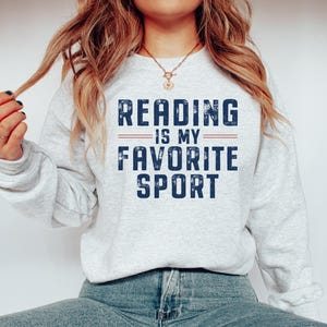 May include: A light gray sweatshirt with the text "READING IS MY FAVORITE SPORT" in distressed navy blue lettering. The text is separated by two horizontal lines. The sweatshirt is paired with blue jeans.