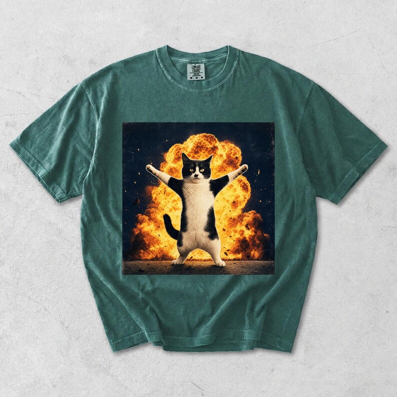Puede incluir: Una camiseta de manga corta de color verde oscuro con un estampado gr&aacute;fico de un gato blanco y negro de pie con los brazos extendidos frente a una gran explosi&oacute;n de color naranja brillante. La camiseta tiene cuello redondo y etiqueta.
