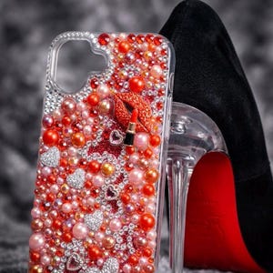 Puede incluir: Una funda transparente para smartphone decorada con una densa disposición de perlas rojas, naranjas y rosas, brillantes de estrás y dijes plateados en forma de corazón. Un pequeño labial tridimensional y un adorno de labios están fijados.